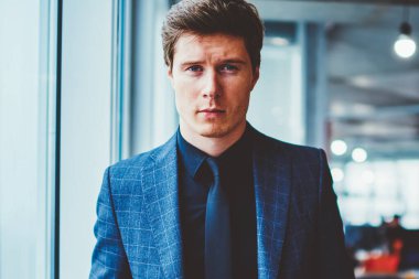 Zarif takım elbiseli ciddi uzman ekonomistin yarı boy portresi ceplerinde, modern şirket ofisinde dikilirken resmi giyinmiş şık, kendine güvenen iş adamı kameraya bakıyor.