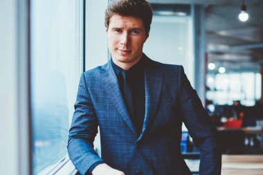 Kendine güvenen şık takım elbiseli bir işadamının portresi ofis penceresinin yanında duruyor ve kameraya bakıyor, yakışıklı beyaz erkek girişimci çalışma alanında vakit geçiriyor ve iş arkadaşını bekliyor.