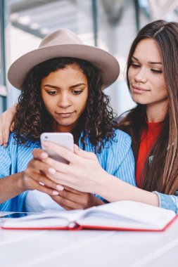 Gündelik giyinmiş çekici hippi kız, yanında oturan ve dijital akıllı telefon cihazıyla e-posta alan kız arkadaşına sarılıyor, cep telefonuyla multimedya uygulaması üzerinden video izleyen büyüleyici kadınlar.