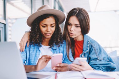 Akıllı telefon cihazları hakkında bilgi paylaşan çekici kadınlar dizüstü bilgisayarla aynı masada oturuyorlar iyi kablosuz bağlantı kullanıyorlar, çok kültürlü hipster kızları cep telefonuyla takipçilerle mesajlaşıyorlar.