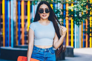 Hobi yaz boş zaman geçirmek için elinde kaykay tutarken kameraya gülümseyen güzel esmer saçlı esmer çekici genç kadın portresi. Teen açık havada duran güneş gözlüğü