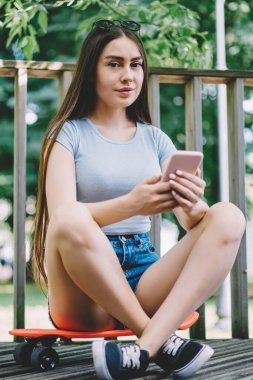 Elinde cep telefonuyla kaykay yaparken kameraya bakan genç pozitif kadın portresi, modern akıllı telefon tutan esmer hippi kız ve açık havada uygulamaları kullanarak sohbet eden kız.