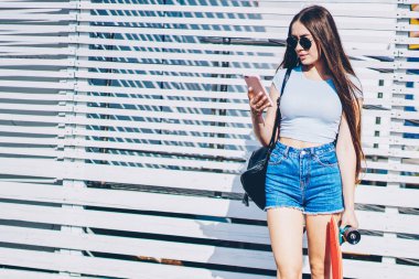 Hippi kız kaykay metin reklam için promosyon arka plan üzerinde ayakta açık havada süre elinde tutan smartphone cep telefonu ile ağdan gelen bildirim okuma rahat kıyafet giymiş