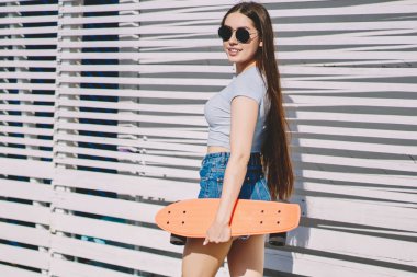 Başarılı bayan patencinin tam boy portresi şık kıyafetler içinde açık havada elinde mini sörf tahtasıyla yürüyor ve güneşli yaz gününün, spor kültürünün ve hipster yaşam tarzının keyfini çıkarıyor.