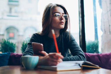 Düşünceli kadın blogcu, kampüs kafeteryasında sosyal ağ ile ilgili ilginç bir fikri olan yeni bir yazı yazmayı planlarken düşünceli bir şekilde kenara bakıyor. Kapalı alanda not defteriyle çalışan düşünceli hippi kız.
