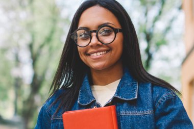 Gülümseyen afro Amerikan kız öğrencinin üniversite bahçesinde açık havada en sevdiğim kitap ile ayakta yarım uzunluk portre. Şık gözlükler ve kot ceket notepad ile yürüme çekici hippi kız