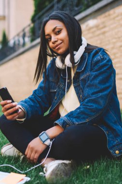 Eğitim ve dinlenme için teknolojisini kullanarak üniversite kampüs çim üzerinde oturan molada dinlenme genç afro Amerikalı kız. Uluslararası öğrenci ödev yaparken cep telefonuna sms gönderme koyu derili