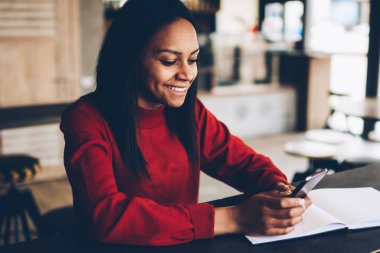 Koyu tenli kız öğrenci kafede oturan e-posta iletisini mutlu gülümseyerek. Başarılı afro Amerikan blogger takipçileri ile sohbet ve paylaşım bilgi ve multimedya cep üzerinde