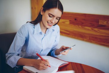 Genç gülümseyen kız öğrenci, kafeteryada arkadaşını beklerken modern akıllı telefon araştırması yaparak ödev yapıyor. 20 'li yaşlardaki neşeli hippi kız cep telefonunu elinde tutuyor ve not defterinde planlama günü düzenliyor.