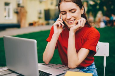 Arkadaşlarıyla akıllı telefondan konuşan ve dışarıda dinlenirken laptopunu kullanan neşeli bayan öğrenci, genç profesyonel serbest çalışan iş görüşmeleri için meslektaşlarını arıyor.