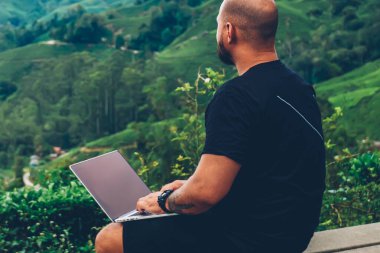 Modern dizüstü bilgisayar için uzaktan çalışmaları sırasında yeşil çiftlik ve yaz gezisi sırasında bitki örtüsü ile yüksek Dağları'nın muhteşem manzarayı ile Hill'de oturan genç adam serbest meslek