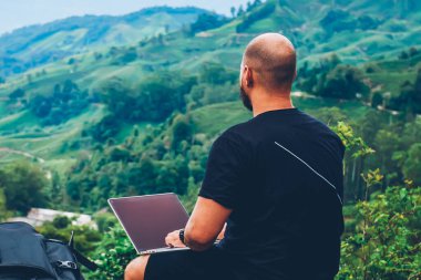 Arkadan erkek turist çalışma serbest treck dağlarda sırasında mesafe işi modern dizüstü bilgisayar, görünüşü. Yeşil manzara ve yüksek tepeler güzellik zevk netbook ile genç adam