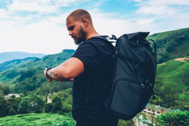 Dalgın sakallı turist Asya dağlar yeşil yerellik içinde yürüyüş Gps izleme için smartwatch bakarak sırt çantası ile. Uzun yürüyüşe çıkan kimse macera trek sırasında kol saati üzerinde uysal yönetme sırt çantası ile