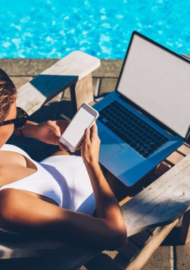 Smartphone ve çalışma serbest içerik için boş kopya alanı ekranlı dizüstü bilgisayar, gelen bildirim okunurken mavi Yüzme Havuzu yakınındaki şezlong üzerinde dinlenme çekici genç kadın