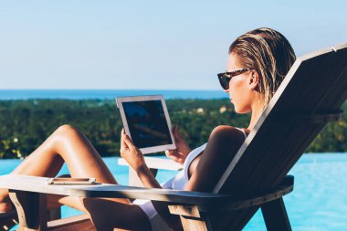 Genç kadın turist dijital dokunmatik yüzey ile sahte alan ve şezlong üzerinde yalan ve havuzda mavi yaz tatili rahatlatıcı sonra güneşlenme Internet üzerinde eğitim izlerken güneş gözlüğü