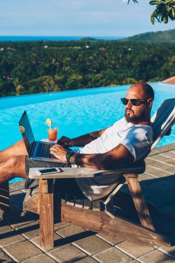 Mavi havuzu muhasebe raporları denetleme ve mesafe yapıyor modern dizüstü bilgisayar iş süre güneş altında güneşlenirken yakınındaki yaz tatili sırasında dinlenme sunlasses genç erkek girişimci sakallı
