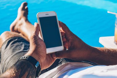 Genç adam turist swinmming havuzu yaz rekreasyon tatil zevk yakınındaki kokteylle şezlong üzerinde yalan smartphone aygıt üzerinde internet sitelerinde bilgi okuma güneş gözlüğü kırpılmış görüntü