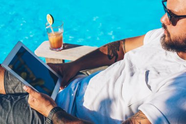 Sakallı işadamı yaz zaman lüks tatil zevk ve tablet sahte mesafe iş yaptığını ve internet kullanımı alanı ile online veritabanı denetimi mavi havuzu yakınındaki şezlong üzerinde güneş gözlüğü