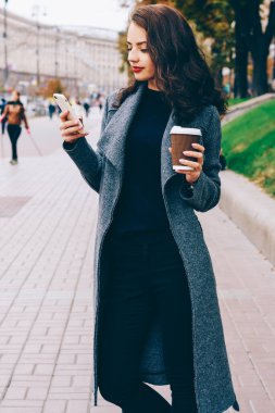 Şık genç kadın ceket elinde tutan ve sohbet online sosyal ağlar üzerinde cep telefonu ayakta Caddesi'ndeki merkezi şehrin trendy ceket giymiş. Reklam için promosyon arka plan