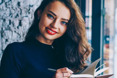 Güzel öğrenci portresi defter elinde tutan ve kameraya gülümseyen ve kahve dükkanında kapalı vintage fotoğraf makinesi ve kitap ile oturma sırasında notlar aşağı yazma trendy gündelik kıyafeti giymiş