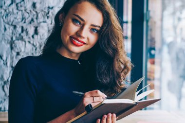 Güzel öğrenci portresi defter elinde tutan ve kameraya gülümseyen ve kahve dükkanında kapalı vintage fotoğraf makinesi ve kitap ile oturma sırasında notlar aşağı yazma trendy gündelik kıyafeti giymiş