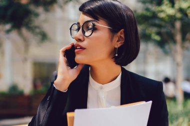 Resmi kıyafeti marka adı veya etiket için kopya alan belgelerle elinde tutan ofis binası yakınındaki bankta otururken cep üzerinde iş ortağı ile konuşmak meşgul kadın Finans Müdürü