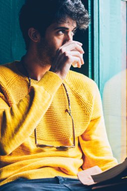Yaratıcı genç adam ise pencereden dışarı bakıyor ve zamanlama oluşturmak için yeni fikirler üzerinde düşünme lezzetli kahve içme sakallı. Düşünceli erkek içecek keyfini rahat sarı Kazak giymiş