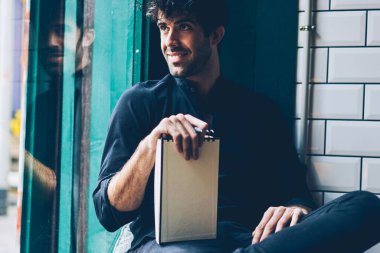 Genç adam gülümseyen alan reklam için Not Defteri ile sahte gidiyor pencereden dışarı bakarak siyah gömlek giymiş. Defter alanı serbest zaman sırasında coworking içinde oturan adamla neşeli hipster