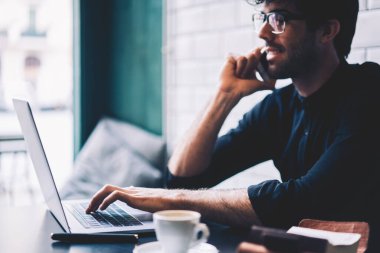 Kafede oturan arkadaşı ile smartphone üzerinde konuşurken bilgi ararken modern dizüstü bilgisayar aygıtının klavye yazarak gözlük neşeli erkek girişimci yan görünüm