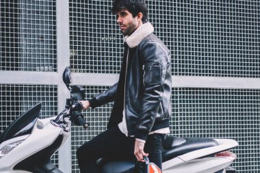 Kentsel ortamda güçlü motor ile kendine güvenen sakallı motorcu deri ceket ayakta beyaz motosiklet yakınındaki giyinmiş. Elinde düz serin motosiklet kırmızı kask ile genç adam