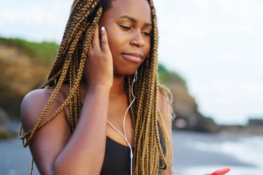 Koyu tenli earhones müzik dinleme genç kadın okyanus tarafından yıkanmış ve spor eğitim için hazırlanıyor plajda durup smartphone bağlı. Afro Amerikan jogging yapan aktif giymek ve el telefon