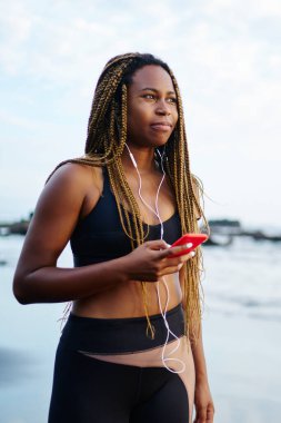 Sportif siyah kadın koşucu sabah sahil üzerinde geziniyor telefon dinleme müzik kulaklık bağlı aktif giyim giymiş. Afro Amerikalı kadın çalıştırmak için eşofman giyen dreads ile