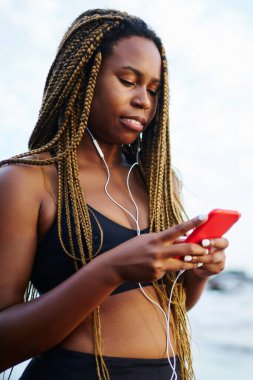 Spor müzik şarkıları sabah deniz kıyısında çalıştırmak için mobil uygulaması seçme dreads ile Afro Amerikan kadın sevgilisi. Egzersiz sırasında smartphone--dan ses dinleme kulaklık jogger koyu derili