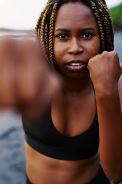Yarım uzunluk portre afro Amerikan kadın boksör kameraya bakarken sahil üzerinde egzersiz sırasında yapıyor. Eğitim sırasında sahilde eliyle aktif giyim boksta koyu tenli sportwoman