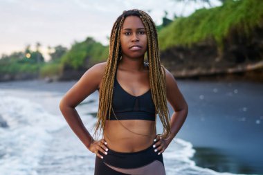 Koyu tenli ve okyanus ile spor sabah eğitimden sonra yıkanmış kıyısında duran güzel dreads ile motive genç kadın portresi. Kameraya bakarak eşofman giymiş Afro Amerikan kadın jogging yapan