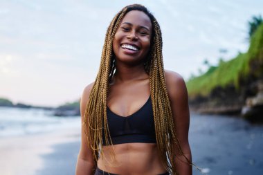 Aktif giyim eğitimden kameraya gülümseyen ise sabah sahil şeridi üzerinde mutlu neşeli afro Amerikalı genç kadın portresi giymiş. Pozitif koyu tenli jogger kıyıda duran spor giyim