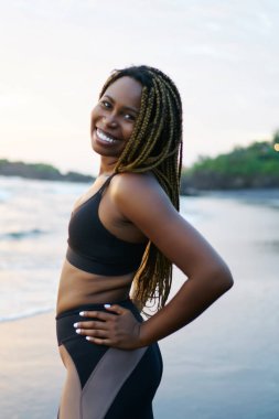 Neşeli afro Amerikan jogger dreads ve deniz kıyısı sabah eğitim sırasında kameraya poz Spor kıyafetleri ile portresi. Aktif giyim ayakta sahil şeridi üzerinde olumlu koyu tenli kadın giyinmiş