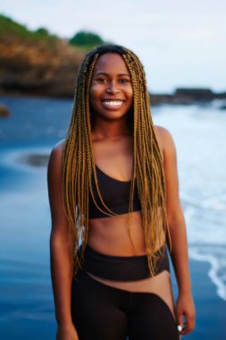 Okyanus su ile yıkanmış kıyısında ayakta dreads ile pozitif afro Amerikan sportwoman portresi. Deniz kıyısı kameraya gülümseyen iş için etkin kıyafetlerini giymiş genç kadın neşeli koyu derili