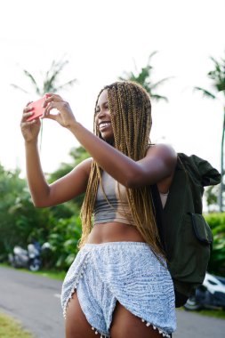 Neşeli afro Amerikan yolcu smartphone aygıtında tropikal yerlesim fotoğrafları yapmak sırt çantası ile. Telefonla yaz gezisi sırasında fotoğraf çekmek dreads ile pozitif koyu tenli turizm