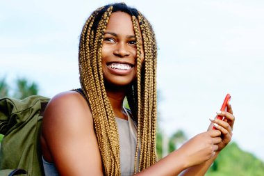 Mutlu afro Amerikalı genç kadın Smartphone'da tropikal ortamda resimlerini yaparken gülüyor sırt çantası ile. Pozitif kadın turist koyu tenli hücresel adada seyahat fotoğraf çekmek