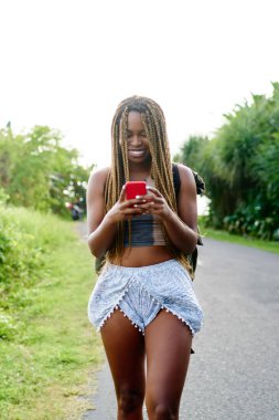 Tropikal terrirory asfalt yol boyunca yaz heyecan verici gezi sırasında geziniyor smartphone aygıt takipçileri ile online sohbet sırasında gülüyor güzel dreads ile pozitif afro Amerikalı turist