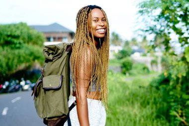 Tropik ada yaz yolculuk sırasında kameraya gülümseyen dreads ile mutlu afro Amerikalı turist portresi. Hafta sonları zevk asfalt yolda geziniyor sırt çantası ile pozitif koyu tenli Gezgin