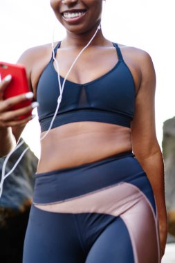 Afro Amerikan kadın jogger kulaklık olarak çalışma için müzik dinleme cep için bağlı eğitim için şık spor elbise giymiş görüntü kırpılmış. Etkin aşınmaya telefon ile yürüyüş sportif kız