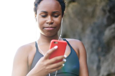 Genç kadın koyu tenli smartphone cep telefonu ile Player'da müzik şarkıları sabah egzersiz açık indiriyor. Afro Amerikan kadın runner etkin aşınmaya sahil eğitim gündoğumu sırasında için ses seçimi