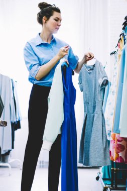 Genç kadın gözlük ile profesyonel stilist moda atelier içinde çalışma ve serin görüntü ile tekstil Giyim Askıları elinde tutan oluşturmak için modaya uygun giysiler seçmek kafa konsantre