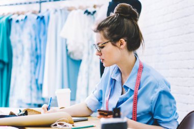 Yaratıcı kadın terzi gözlük ve ekipman ve modern latop bilgisayar kendi şık atelier içinde okul sırası vasıl oturan setches çizim yeni giysi tasarımı teyp yan görünüm