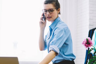 Showroom dikiş masada oturan telefon üzerinde çalışma programı organize Yöneticisi. Yaratıcı terzi koleksiyonu moda sanayi olay için hazırlanması için tasarımcı ile smartphone üzerinde konuşurken