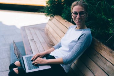 Çevrimiçi kurslardan memnun olan neşeli kız öğrencinin portresi açık havada netbook yoluyla öğreniyor, serbest çalışan mutlu hippi kız dizüstü bilgisayarında dizüstü bilgisayarda çalışıyor, bankta oturuyor.