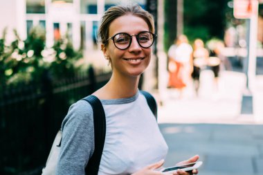 Şehir merkezinde gezen mutlu kadın turistin portresi. Uygulama yoluyla navigasyon için akıllı telefonu tutuyor. Neşeli hippi kız kameraya bakıyor.