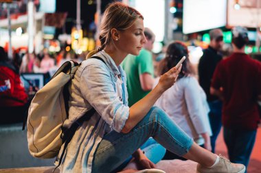 Genç kadın kadın Gezgin kalabalık Times Square'de akşam oturan smartphone üzerinden blog resimleri ilanıyla New York'a yolculuk sırasında dolaşım Internet kullanarak cep telefonunda mesaj okuma